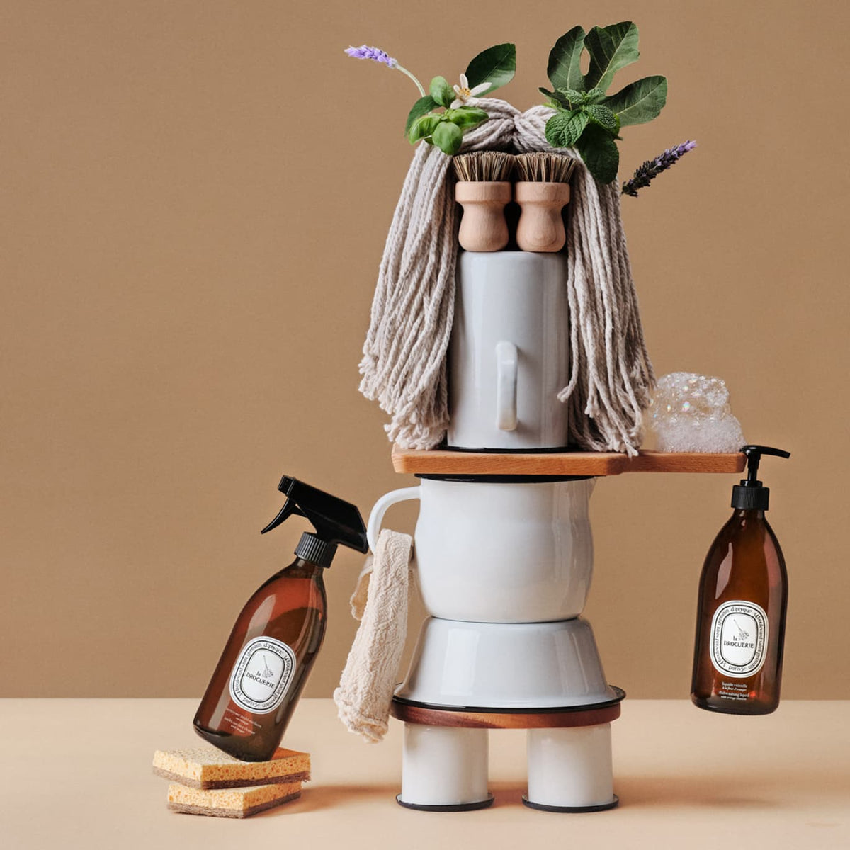 Diptyque La Droguerie, Reinigungssprays und Mopp zu einer Figur arrangiert vor beigem Grund