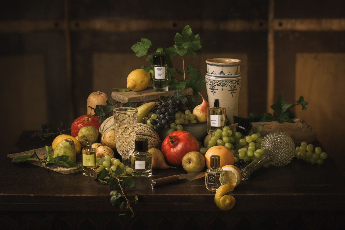 Duft und Kultur Editorial – Früchte der Versuchung, Stillleben mit Parfumflakons zwischen Granatäpfeln, Trauben, Birnen und Zitronen