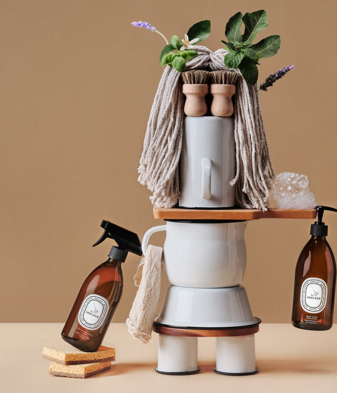 Diptyque La Droguerie, Reinigungssprays und Mopp zu einer Figur arrangiert vor beigem Grund